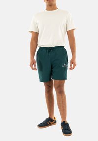 Calções de banho verdes com cintura elástica e cordão, apresentando bolsos laterais e um pequeno logótipo. Usados com uma t-shirt leve e ténis.