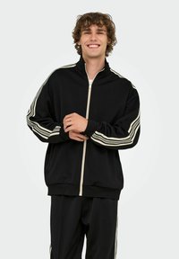 Schwarze Sportjacke mit Reißverschluss, weißen Streifen an den Ärmeln, gerippten Bündchen und Stehkragen. Weicher, glatter Stoff.