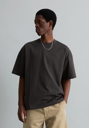 Young Black man in loose black t-shirt and beige pants stands with one hand in pocket, looking to his left against a plain background.