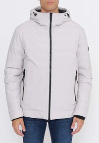 Uomo che indossa una giacca con cappuccio grigio chiaro con cerniera nera e tasche con cerniera, abbinata a jeans blu, mostrato dal collo all'anca.