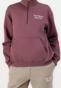 Purple, zip-up sweatshirt with high neck, front pocket, and white "New Balance Athletic Club" logo; paired with beige sweatpants.