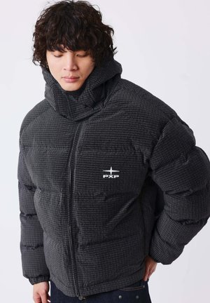Joven con cabello rizado que lleva una chaqueta acolchada oscura con un cuello alto y el logo "PXP", mirando hacia abajo con una mano en la cadera.