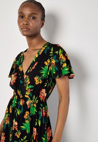Blomstret kjole med sort baggrund, der har en dyb V-udskæring, korte flæseærmer og en elastisk talje. Farverne inkluderer orange, grøn og gul.