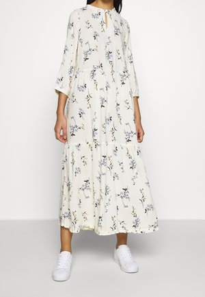 Mujer vistiendo un vestido largo blanco con flores, mangas tres cuartos y zapatillas blancas, de pie frente a un fondo blanco.