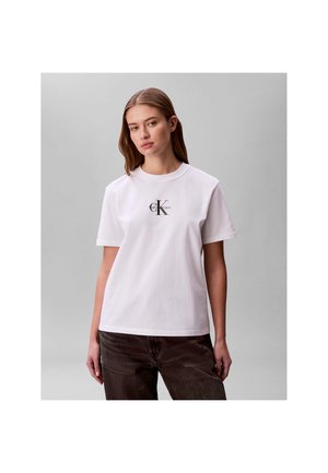 Giovane donna con t-shirt bianca Calvin Klein e pantaloni scuri, in piedi davanti a uno sfondo grigio uniforme con espressione neutra.