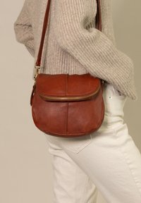 Borsa a tracolla in pelle marrone di forma circolare, con un cappuccio frontale e dettaglio in zip dorata, indossata da un modello con jeans chiari.