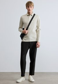 Μακρυμάνικο πουλόβερ πόλο σε ανοιχτό μπεζ χρώμα με γιακά και κεντημένο λογότυπο, συνδυασμένο με μαύρο παντελόνι στενής γραμμής και λευκά αθλητικά παπούτσια. Κρατάει μια μαύρη τσάντα.