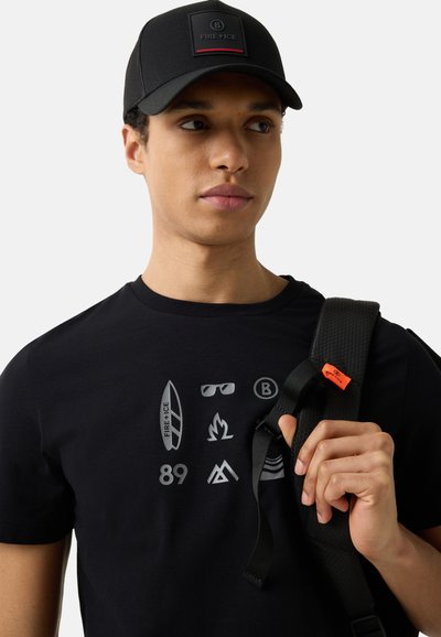 Mladý muž s černou čepicí s nápisem "B FIRE + ICE", černým tričkem s potiskem symbolů, držící popruh černého batohu s oranžovou visačkou.