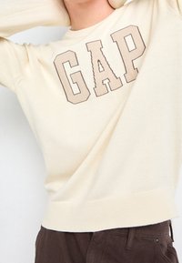 Person som bär en krämfärgad GAP-tröja med stora beige bokstäver, ihop med mörkbruna byxor, lyfter armarna bakom huvudet.