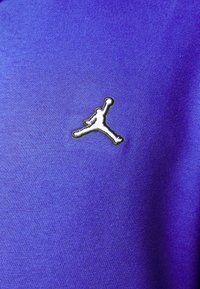 Fialová látka s hladkou strukturou ukazuje bílé vyšívané logo Jumpman v dolním rohu. Design je minimalistický a moderní.