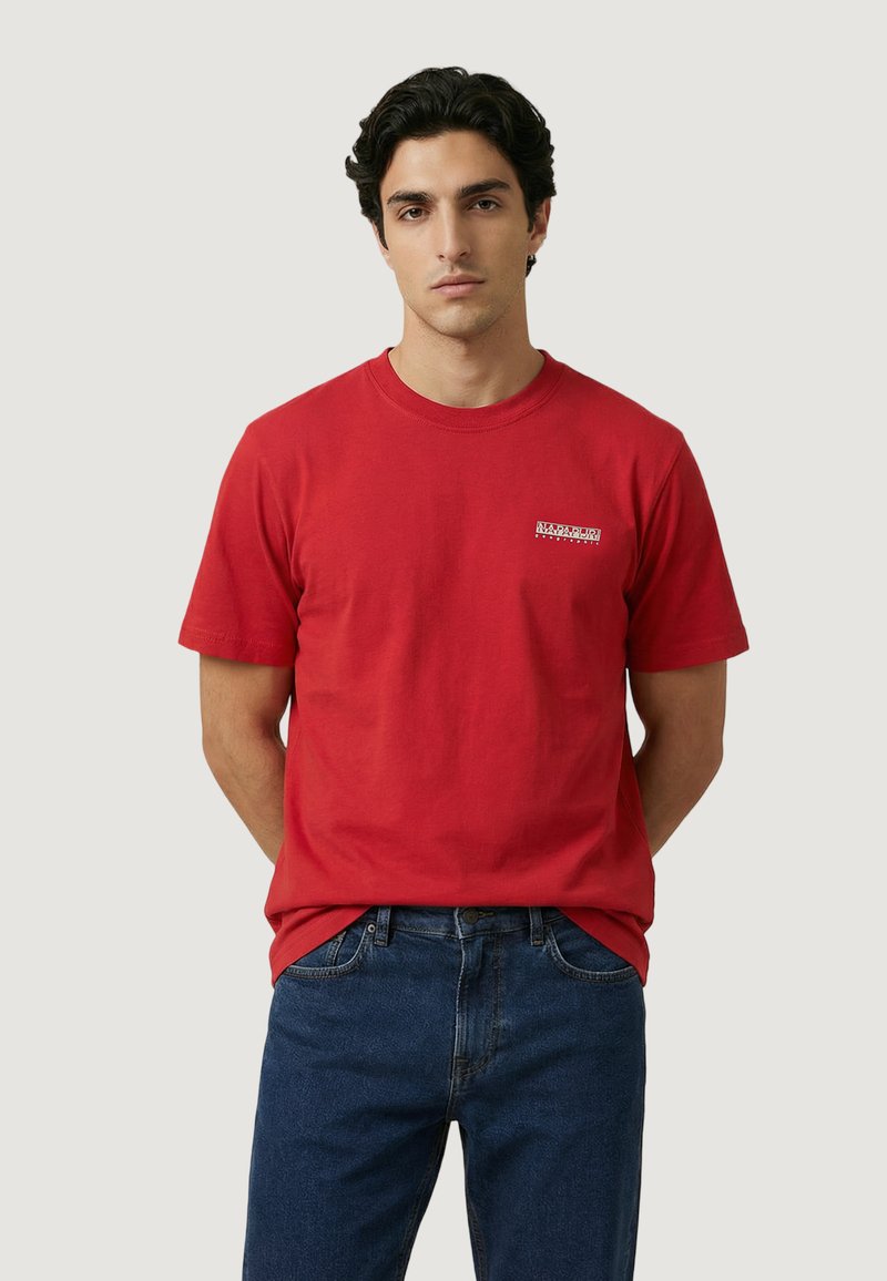 Giovane uomo che indossa una maglietta rossa a maniche corte con un piccolo logo bianco sul petto, abbinata a jeans blu, in piedi con le mani dietro la schiena.