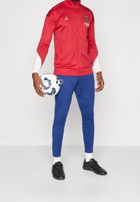 Chaqueta roja con cremallera y detalles blancos y logo, combinada con pantalones ajustados azules y zapatos de fútbol negros, sosteniendo un balón de fútbol con un estampado.