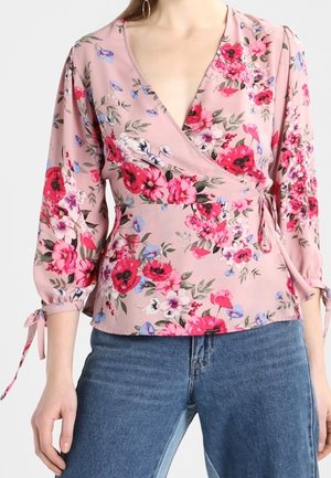 Blusa cruzada rosa con estampado floral en rojo, blanco y azul, mangas tres cuartos con lazos, combinada con vaqueros azules.
