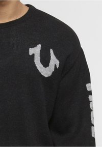 Black sweater with a textured finish. Features a grey logo on the chest and white text along the sleeve. Round neckline and relaxed fit.