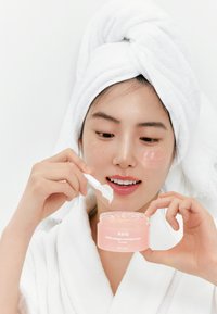 Maschera notturna in gel di collagene rosa in un barattolo trasparente, tenuto in mano con una spatola bianca. La persona indossa un asciugamano bianco e un accappatoio. Sfondo semplice.