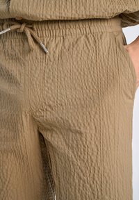 Pantalones de mezcla de algodón en marrón suave con un patrón de cuadrícula texturizado. Presentan una cintura elástica con cordón y bolsillos laterales.