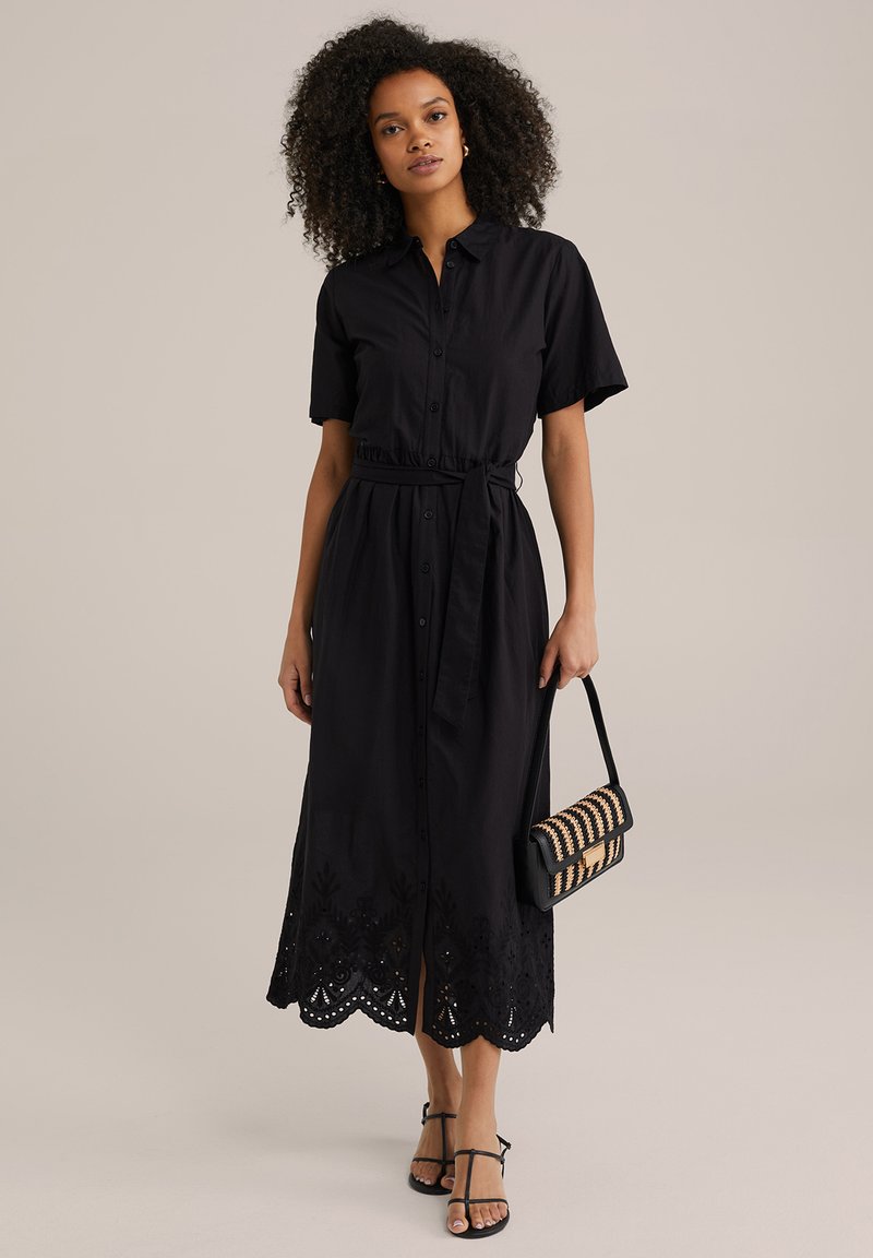 Abito camicia nero con bottoni, maniche corte, vita cinte e orlo ricamato. Abbinato a una borsa a tracolla a righe e sandali con lacci.
