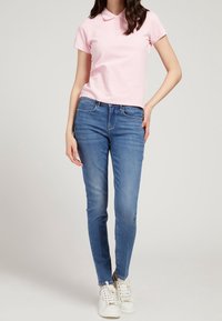 Camisa polo rosa claro de manga corta, combinada con jeans ajustados azules y zapatillas blancas, destacando el diseño y el ajuste de la vestimenta casual.
