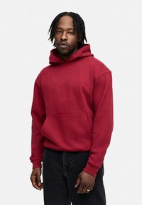 Roter Baumwoll-Hoodie mit einer Kängurutasche vorne, lockerer Passform und gerippten Bündchen. Getragen mit dunklen Jeans.