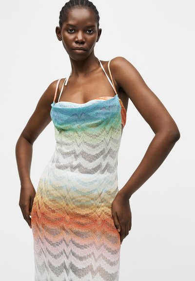 Νεαρή γυναίκα που φοράει αμάνικο φόρεμα με πολύχρωμο διαβαθμισμένο σχέδιο και λεπτές τιράντες, στέκεται με τα χέρια στη μέση μπροστά σε απλό φόντο.