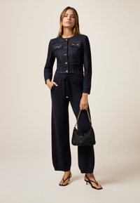 Tumman denim-kangas cropped-takki, jossa on nappikiinnitys ja kaksi rintataskua, yhdistettynä korkeavyötäröisiin tummansinisiin housuihin ja pieneen mustaan käsilaukkuun, jossa on ketjuolkaimet.