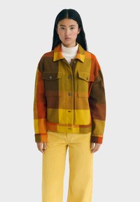 Chaqueta de cuadros con cuello en tonos naranja, amarillo, marrón y verde; cuenta con dos bolsillos en el pecho y cierre de botones. Llevada sobre un cuello de tortuga blanco.