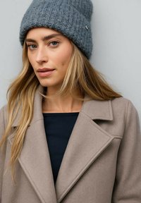 Gorro de punto gris con textura acanalada y un adorno metálico. Abrigo beige con solapas de muesca, llevado sobre una blusa azul marino.