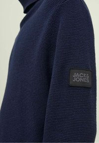Maglione a maglia testurizzata di un blu navy scuro con una toppa rettangolare nera sulla manica che mostra la scritta "JACK & JONES" in lettere grigie.