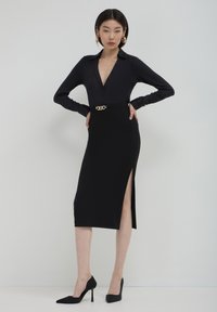 Blouse noire à manches longues avec boutons, associée à une jupe crayon avec fente latérale et accent en chaîne dorée à la taille, complétée par des talons noirs.