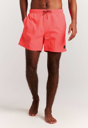 Homme debout pieds nus portant un short de bain corail avec un cordon noir et un petit logo rond noir sur la jambe gauche, sur fond blanc.