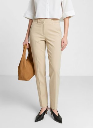 Personne portant un pantalon beige sur mesure, une chemise blanche courte, des talons pointus noirs, et tenant un sac à main en cuir marron à ses côtés.