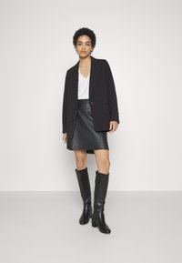 Blazer noir ajusté, haut en V blanc, jupe en cuir noir et bottes noires à talons mi-cuisses. Textures lisses et design structuré.