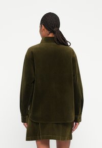 Oliven grøn kordfløjlsskjorte med lange ærmer, knapmanchetter og en delt kant. Teksturen er ribbet, hvilket fremhæver dens taktile detalje.