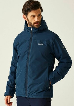 Mężczyzna ubrany w granatową, wodoodporną kurtkę Regatta z kapturem, zapinaną na zamek, stojący z jedną ręką w kieszeni, patrzący w bok.