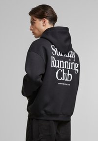 Felpa nera con cappuccio in misto cotone, caratterizzata da grandi scritte bianche "Sunday Running Club" sul retro e tessuto testurizzato.