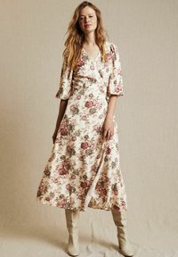 Kukkakuosiinen kietaisumekko vaaleassa sävyssä, jossa on vaaleanpunaisia ja vihreitä ruusuja. Mekossa on kyynärpituisten hihansuiden lisäksi V-pääntie ja virtaava siluetti. Ivory-väriset nilkkakengät.