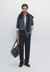 Camicia blu a maniche lunghe sopra una maglietta bianca, pantaloni scuri, borsa in pelle nera e scarpe scure, con un maglione marrone drappeggiato sulle spalle.