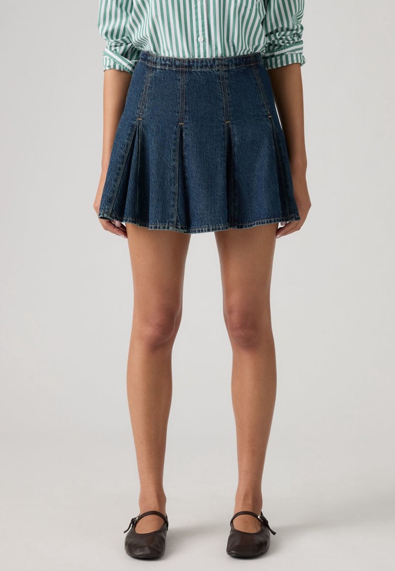 Femme portant une mini-jupe plissée en denim foncé, une chemise rayée verte et blanche aux manches retroussées, et des chaussures Mary Jane noires.
