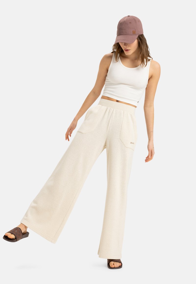 Crèmekleurige wijde broek met een zachte textuur en twee voorkruiden, gedragen met een aansluitend wit hemd en een lichtroze pet.