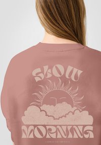 Roze sweatshirt met een grote afbeelding met "SLOW MORNING," zon en wolken in een gedempte crèmekleur, met een textuurafwerking.