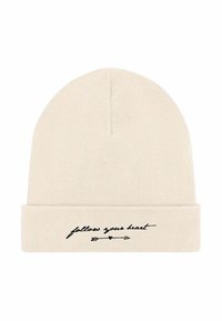 Cappellino a maglia in beige chiaro con risvolto. Presenta un testo corsivo nero "segui il tuo cuore" e una grafica con una freccia. Tessuto morbido.