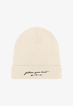 Cappellino a maglia in beige chiaro con risvolto. Presenta un testo corsivo nero "segui il tuo cuore" e una grafica con una freccia. Tessuto morbido.