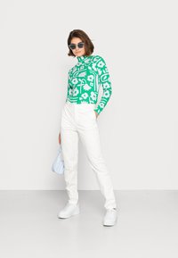 Blusa verde de manga longa com padrão floral branco, combinada com calças brancas e ténis brancos. Modelo segura uma bolsa azul-clara.