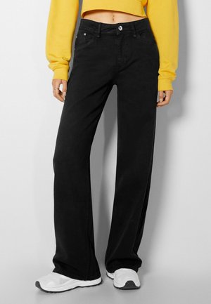 Osoba ubrana w czarne dżinsy szerokie, żółty krótki sweter i białe trampki, stojąca na tle jednolitego jasnego tła.