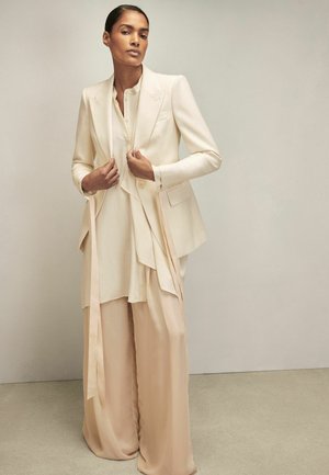 Vrouw draagt een crème tailored blazer, bijpassende gelaagde blouse en beige broek met wijde pijpen, staand tegen een effen neutrale achtergrond.