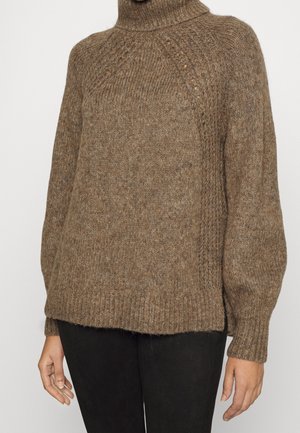 Pull à col roulé marron en maille avec des motifs texturés sur la poitrine et les manches, porté avec un pantalon noir.
