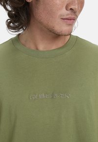 Olivgrünes Baumwoll-T-Shirt mit Rundhalsausschnitt und dezentem, in einer helleren Farbe gesticktem "Quiksilver"-Logo auf der Vorderseite. Weiche Textur.