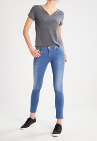 Woman wearing a short-sleeve gray V-neck shirt, light blue skinny jeans, and black sneakers standing with one hand in pocket.