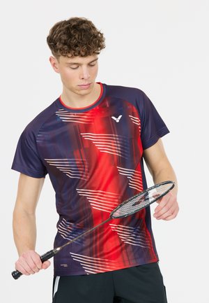 Ung mand i en rød og marineblå sportsskjorte holder og inspicerer en badmintonketcher mod en ensfarvet baggrund.