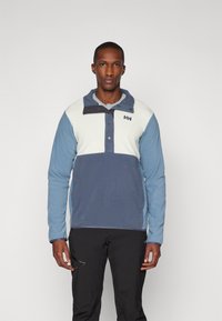 Pullover leggero con un design a blocchi di colore blu e bianco, dotato di chiusura a bottoni a pressione, alto colletto e morbida texture del tessuto.